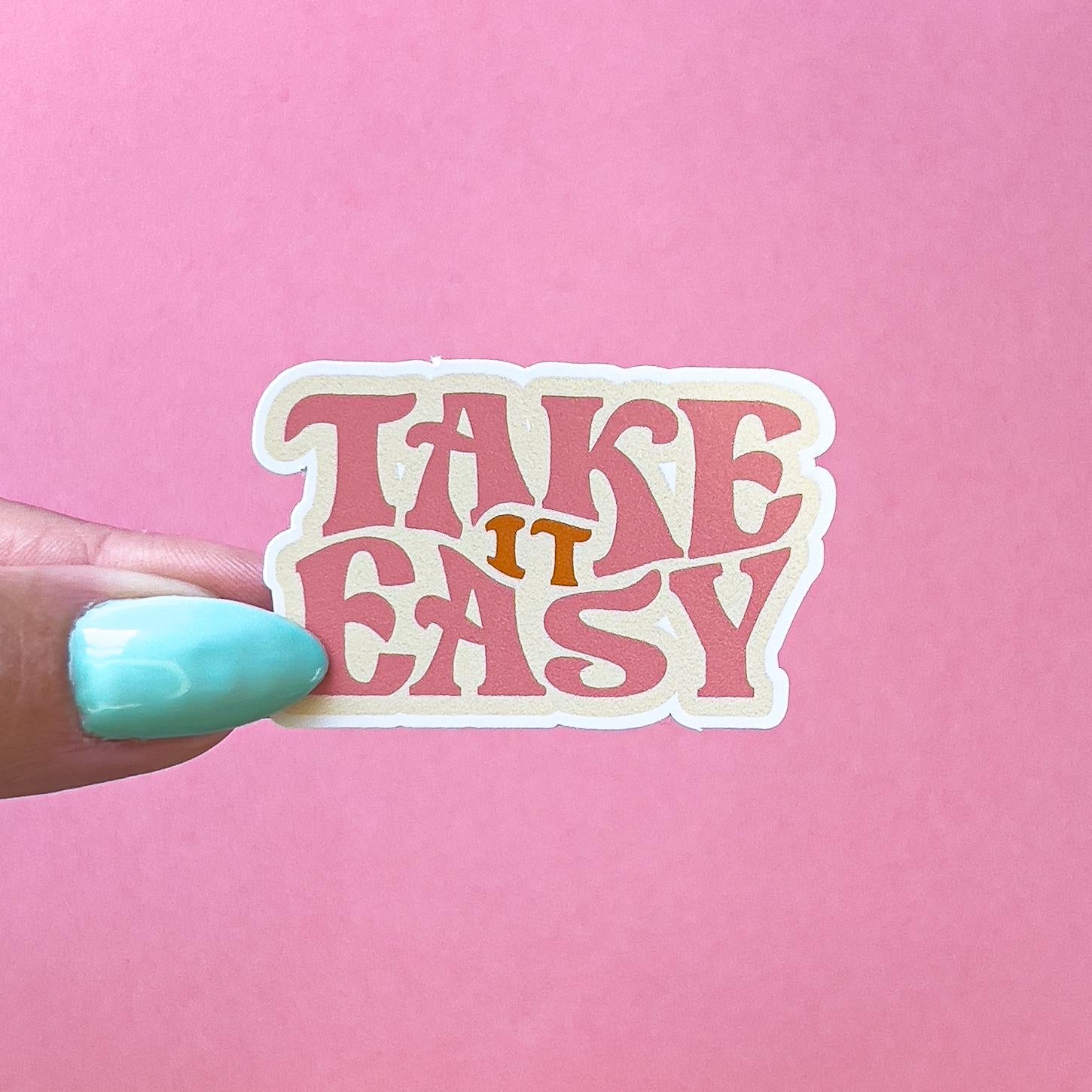 A vinyl decal sticker with the text 'Take it easy' in a retro font. The text is pink with a white outline and the word 'it' is in orange.