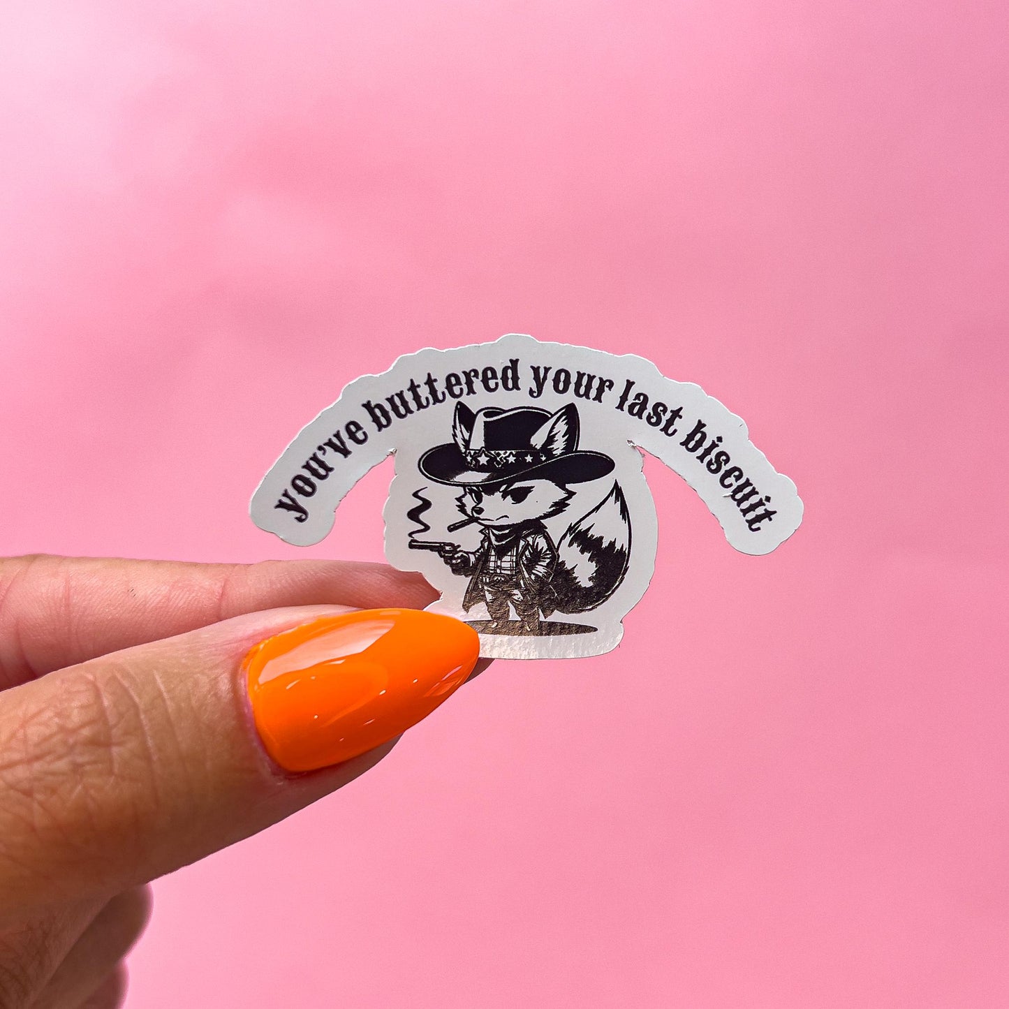 A vinyl decal sticker of a black and white cartoon fox smoking a cigar while wearing a cowboy hat and holding a gun. The text on the sticker reads 'You've buttered your last biscuit.'