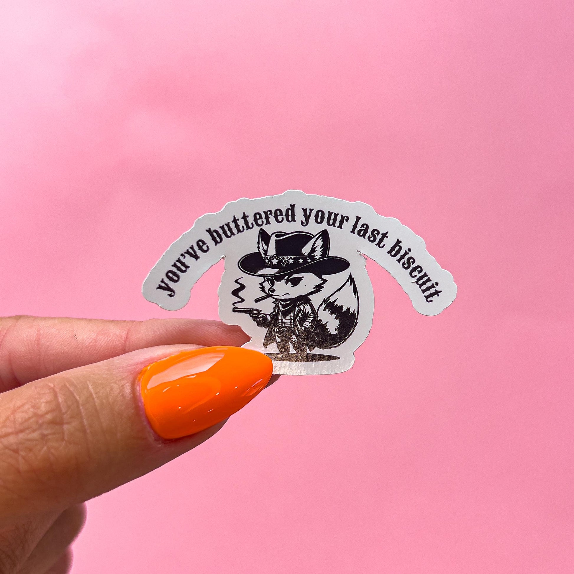 A vinyl decal sticker of a black and white cartoon fox smoking a cigar while wearing a cowboy hat and holding a gun. The text on the sticker reads 'You've buttered your last biscuit.'