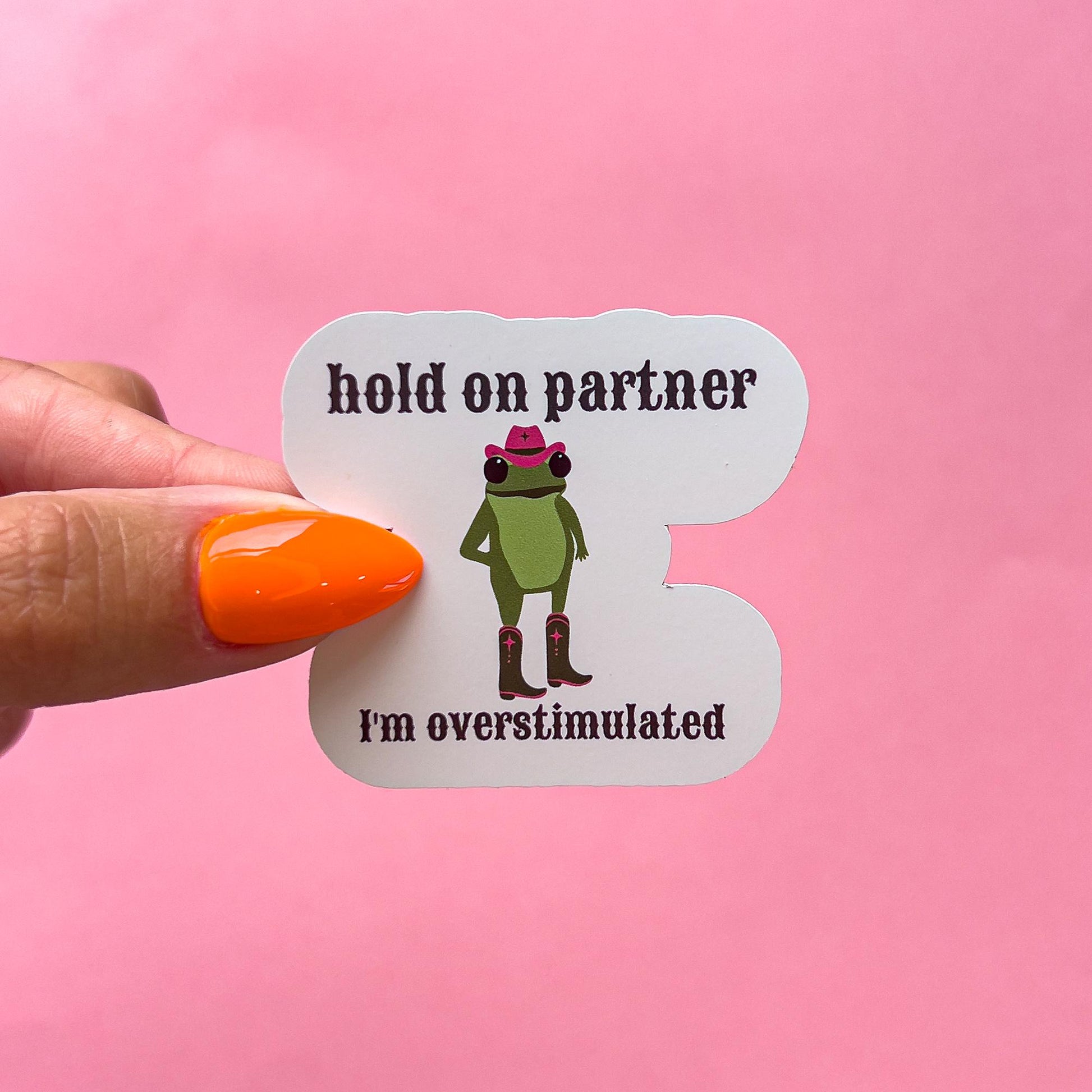 A vinyl decal sticker of a cartoon cowboy frog. The frog is green and wearing a pink cowboy hat and brown cowboy boots. The frog is standing awkwardly, slightly turned to one side. The text reads "hold on partner I'm overstimulated" in western font.
