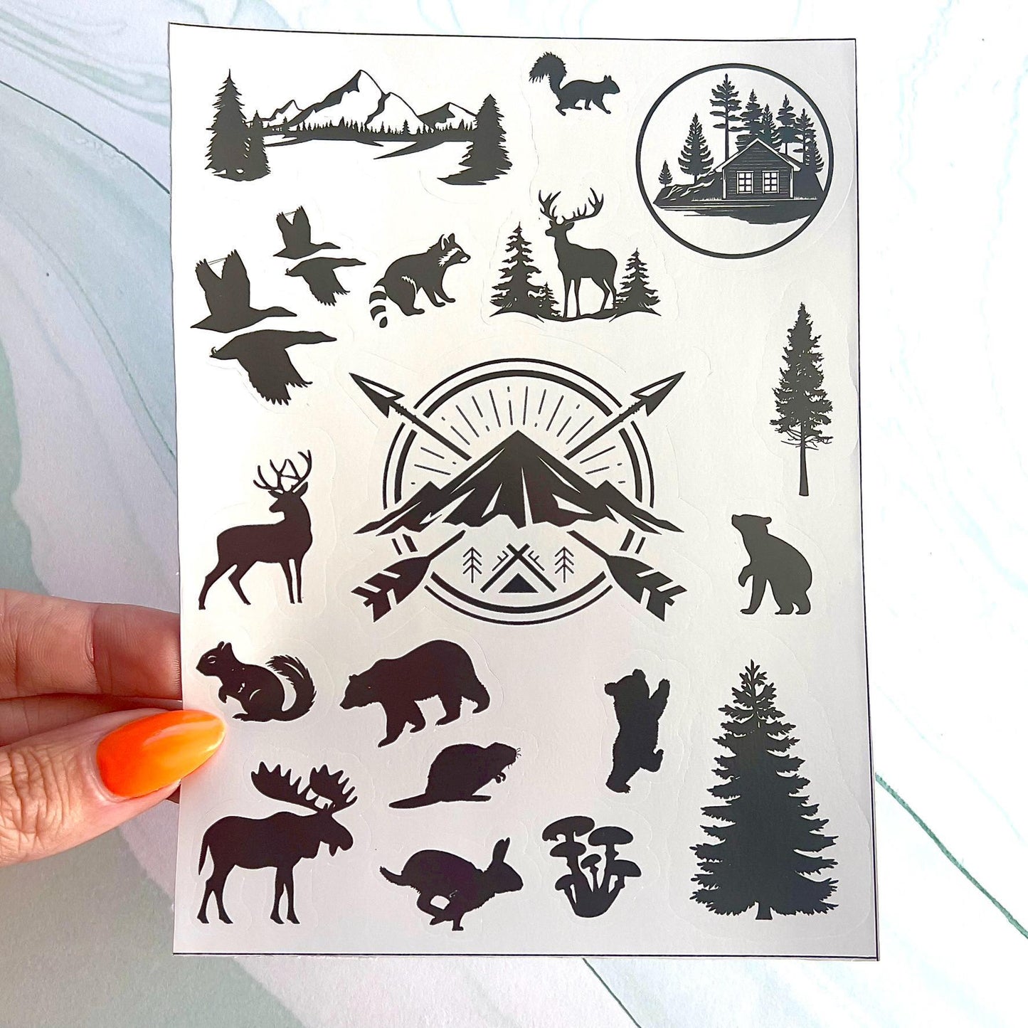 A sheet of black and white stickers featuring a variety of nature-themed designs, including a mountain range, a cabin, a deer, a bear, a squirrel, a moose, a rabbit, a tree, and a bird. The stickers are on a white background.