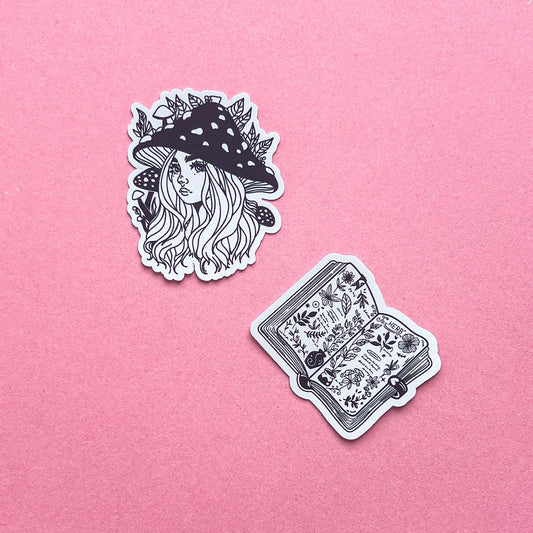 Two black and white vinyl decal stickers. One is of a cartoon style girl/woman with long hair, big eyes, a small upturned nose. She is wearing a mushroom hat with mushrooms and leaves around her. The other sticker is an open spell book. The pages feature botanical pictures and words.