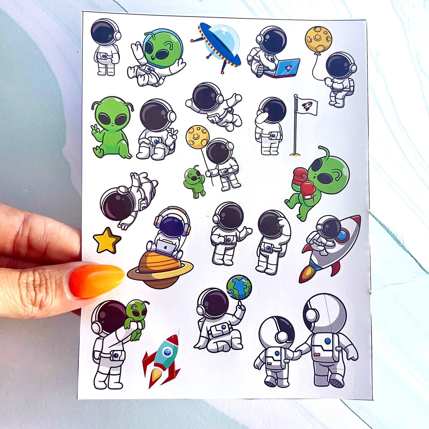 A sheet of stickers featuring cartoon astronauts and aliens. The stickers feature a variety of designs, including astronauts in spacesuits, aliens with green skin and antennas, and spaceships. The stickers are perfect for adding a fun and whimsical touch to any project.