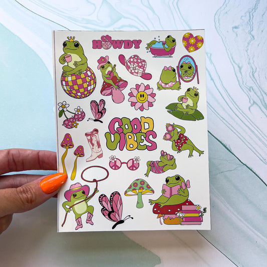 A sheet of stickers featuring frogs in various outfits and poses. The stickers include a cowboy frog with a lasso, a frog on a stack of books with a bow, frogs in swimsuits and heart shaped sunglasses, a frog wearing a crown while sitting on a disco ball, a cowboy frog playing guitar , a pink cowboy hat, butterflies, mushroom, cowboy boots, "GOOD VIBES", "HOWDY" with a cowboy hat on a disco ball instead of an "O", ect. The stickers are shades green, pink, and yellow.