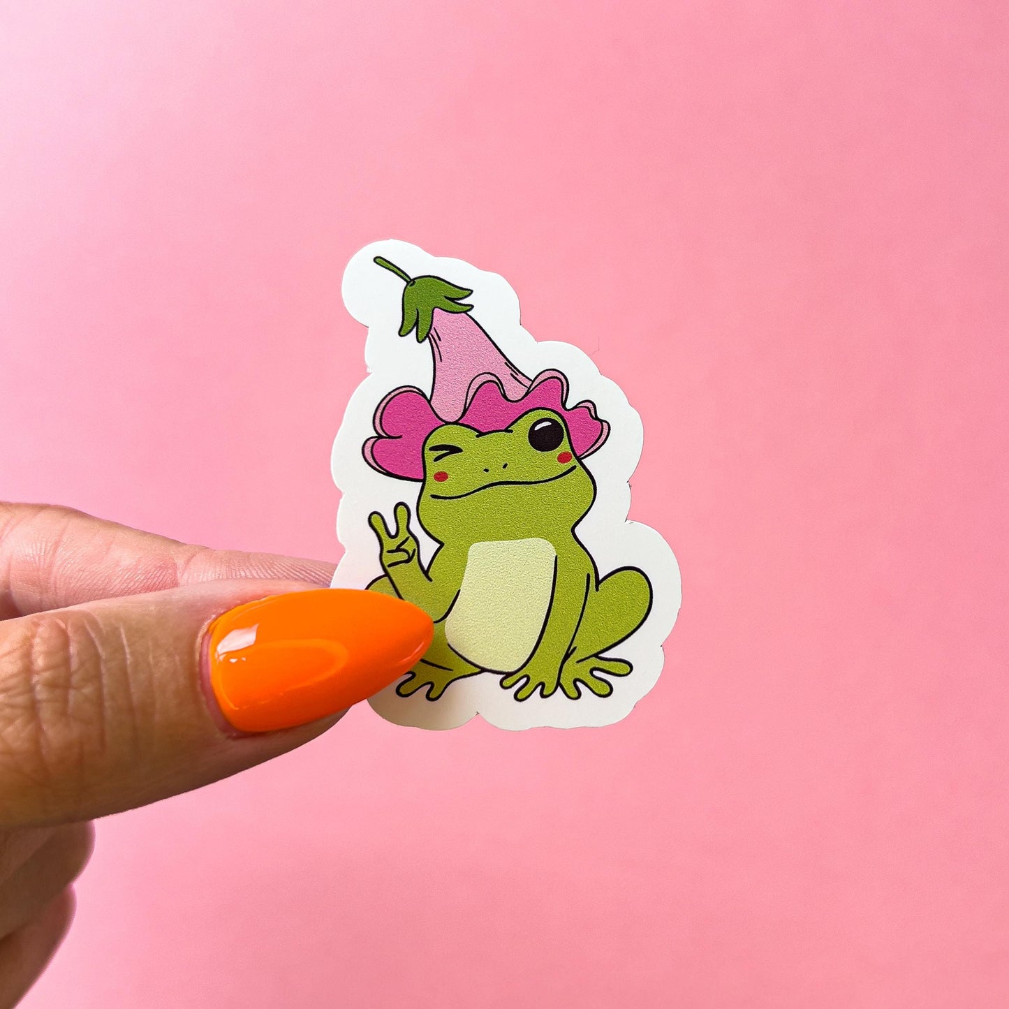 A vinyl decal sticker of a green, cartoon frog with blushed cheeks and holding up a peace sign. The frog is wearing a pink flower as a hat.